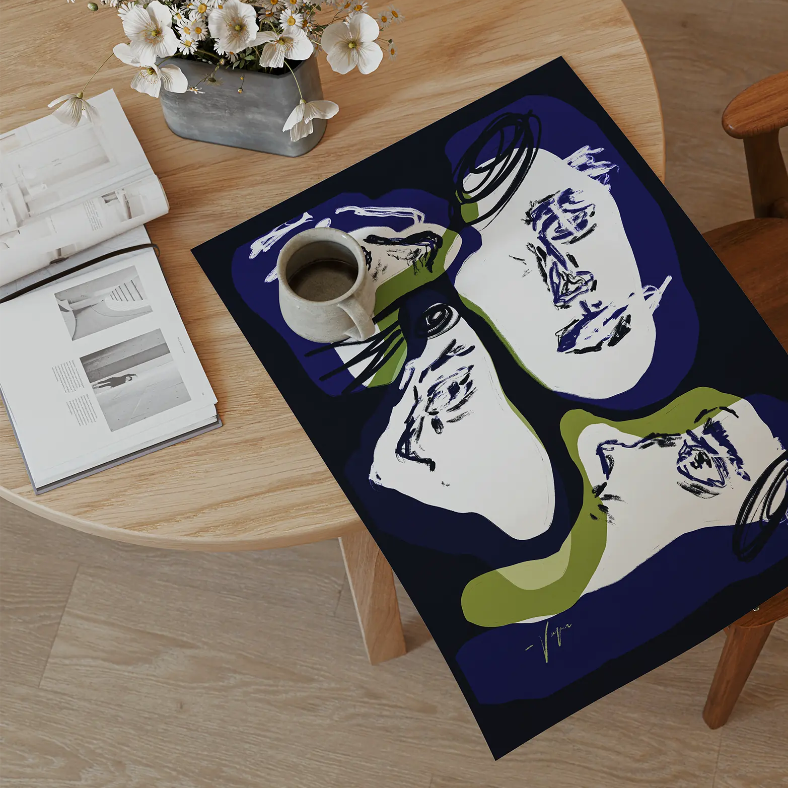A rectangular art print or poster featuring an abstract face design in navy, white, and olive green, laid on a light wood floor next to a wooden chair and a round wooden table with books and flowers.