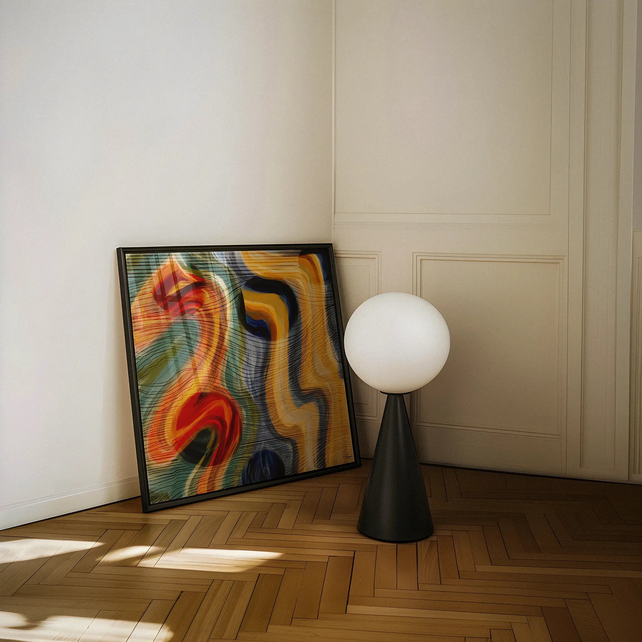 A framed abstract wall art print with swirling red, orange, yellow, blue, and green tones leans against a cream paneled wall on a wood herringbone floor, next to a black table lamp with a white globe shade.