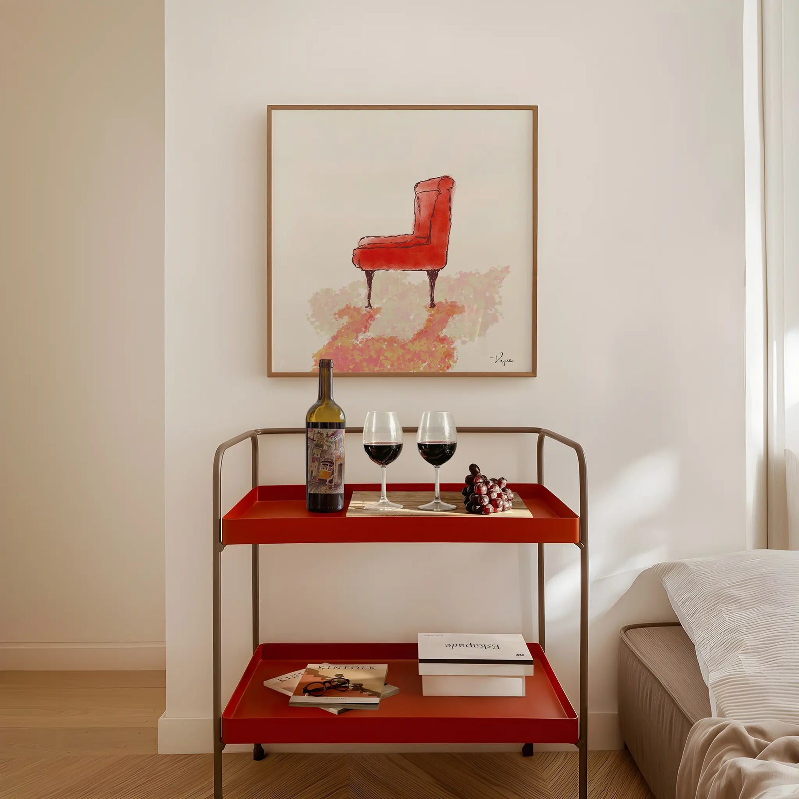 A two-tier red metal and wood serving cart styled with a wine bottle, two wine glasses, grapes, and books, set against a warm beige wall with a framed red chair painting above.