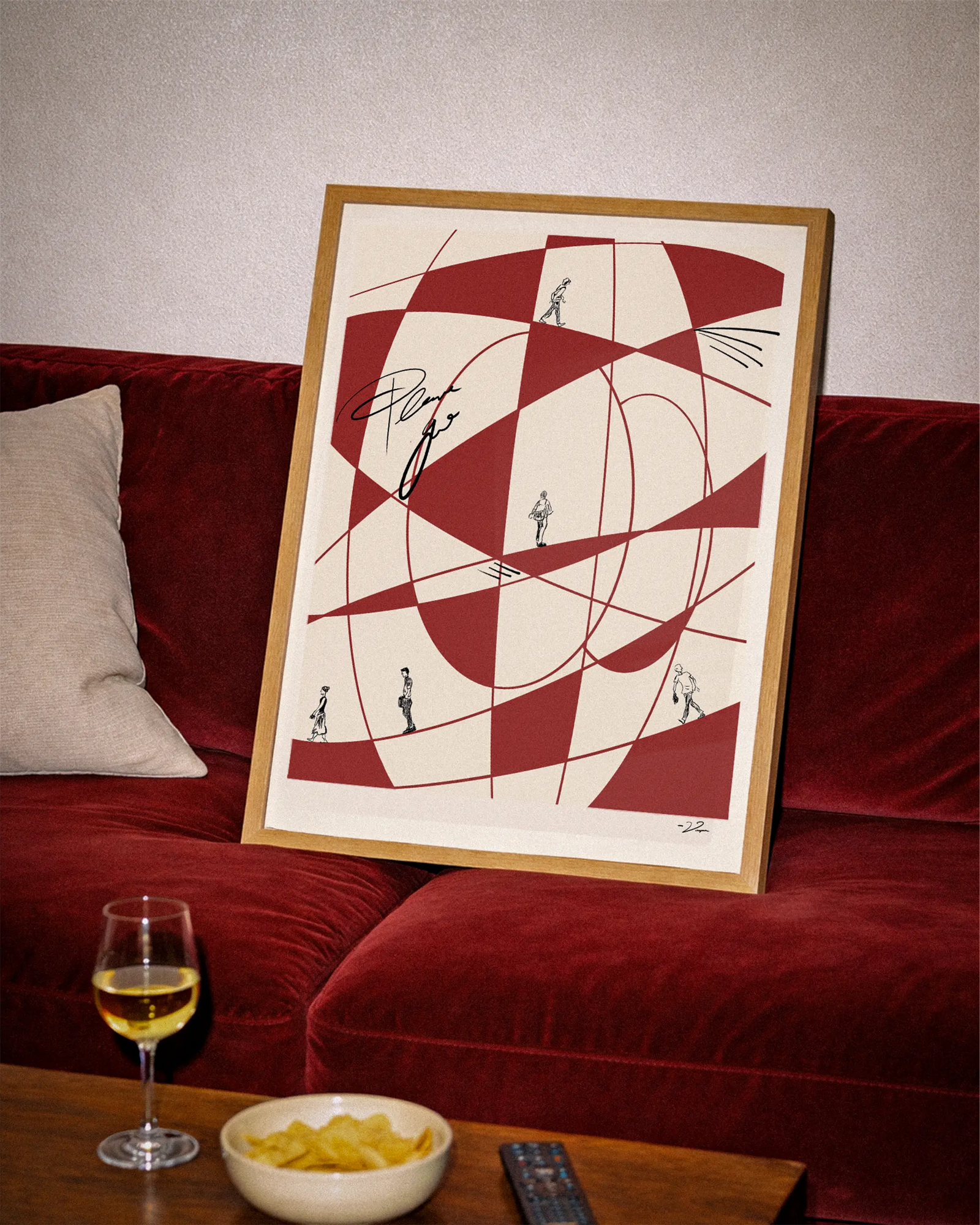 A framed abstract art print with red and cream geometric shapes and small black figures, leaning against a red velvet sofa in a living room setting with a wine glass and bowl of chips on a wooden table.