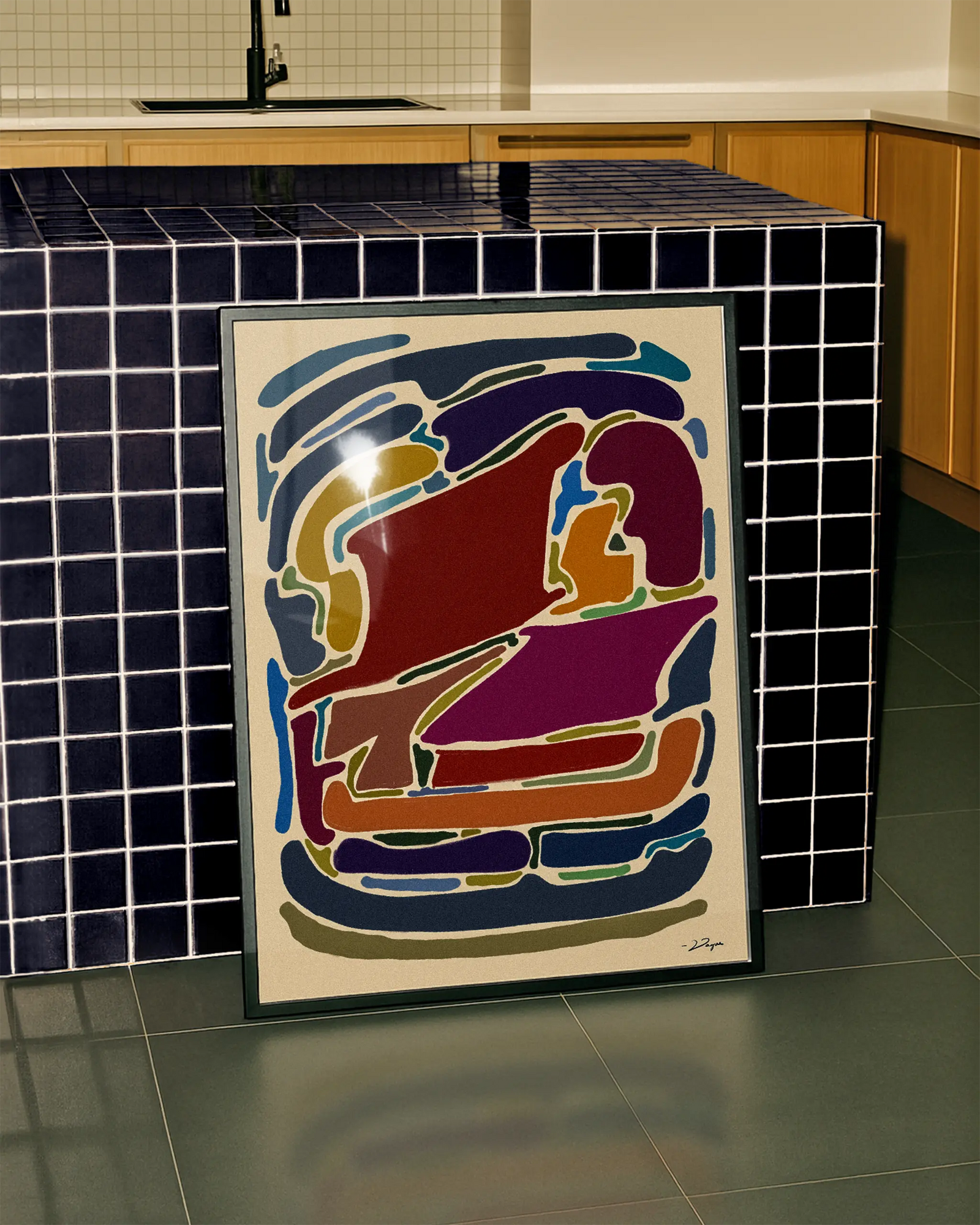 A framed abstract art print with colorful organic shapes in red, purple, blue, orange, and yellow on a cream background, leaning against a dark blue tiled kitchen island.
